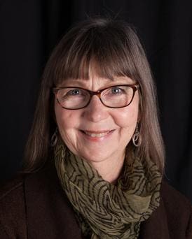 00f185b01cd1328a117e4fe31543d888 A smiling woman with glasses, wearing a scarf and a brown jacket against a dark background.